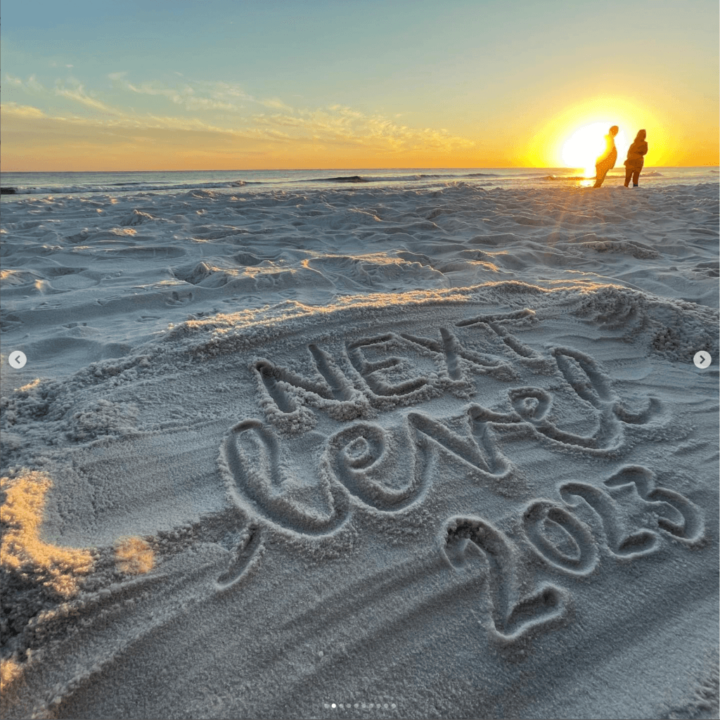 The image shows a sandy beach at sunset, with "NEXT LEVEL 2023" written in the sand. Two silhouetted people stand near the water's edge under the sun.
