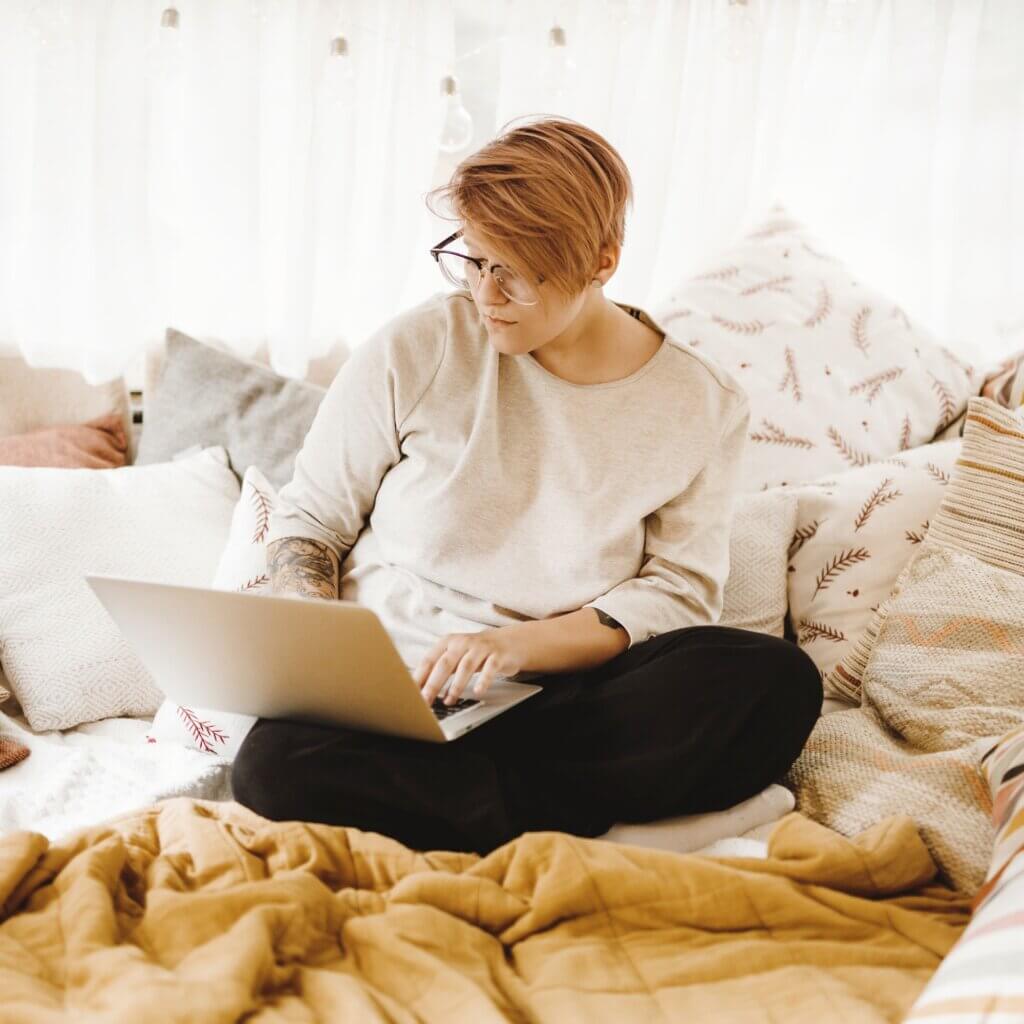 A person with glasses and a tattoo sits on a bed covered with pillows and a mustard blanket, working on a laptop in a cozy room.