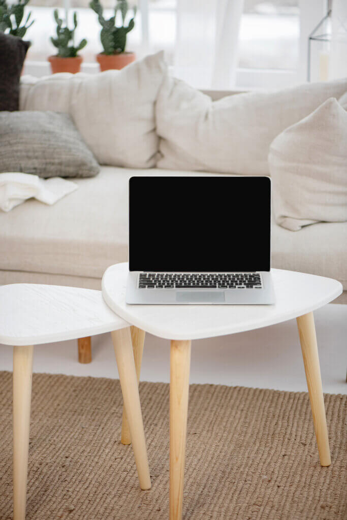 An open laptop with a black screen sits on a white round table with beige wooden legs, placed on a textured rug, near a couch with pillows.