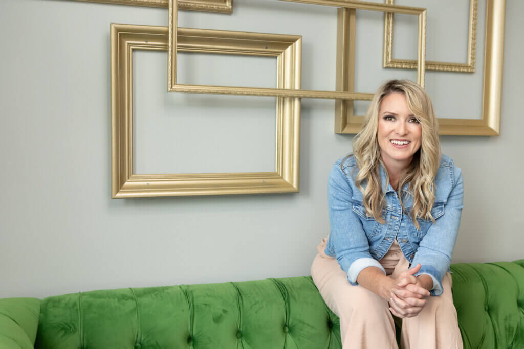 A smiling person sits on a green tufted sofa, wearing a denim jacket. Gold frames on a light blue wall form decorative empty rectangles.