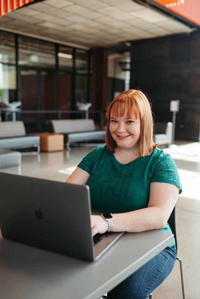 A person with red hair is smiling and using a laptop at a table in a modern office setting with large windows and lounge areas.