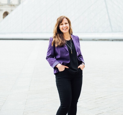 A person is smiling at the camera, wearing a purple blazer and black pants, standing in front of a blurred geometric structure with hands in pockets.