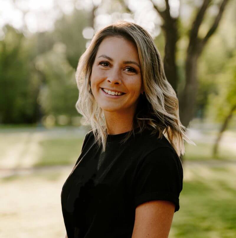 A smiling person with blonde hair, wearing a black top, stands outdoors with trees in the blurred background during the daytime.