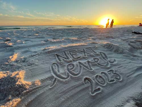 This image shows a beach at sunset with "Next Level 2023" written in the sand. Silhouettes of three individuals stand in the background near the water.