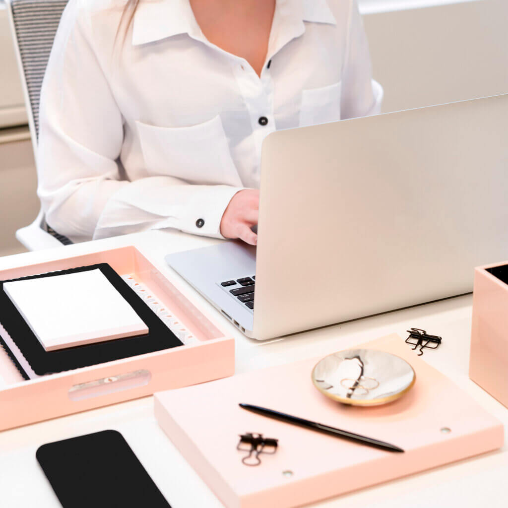 A person is working on a laptop at a neat desk with pink stationery, including notebooks, a pen, a small dish, and paper clips.