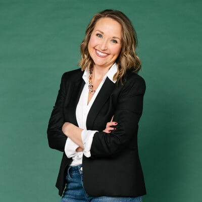 A person stands against a green background, smiling, with arms crossed. They wear a black blazer, white top, and denim jeans, projecting a professional yet relaxed demeanor.