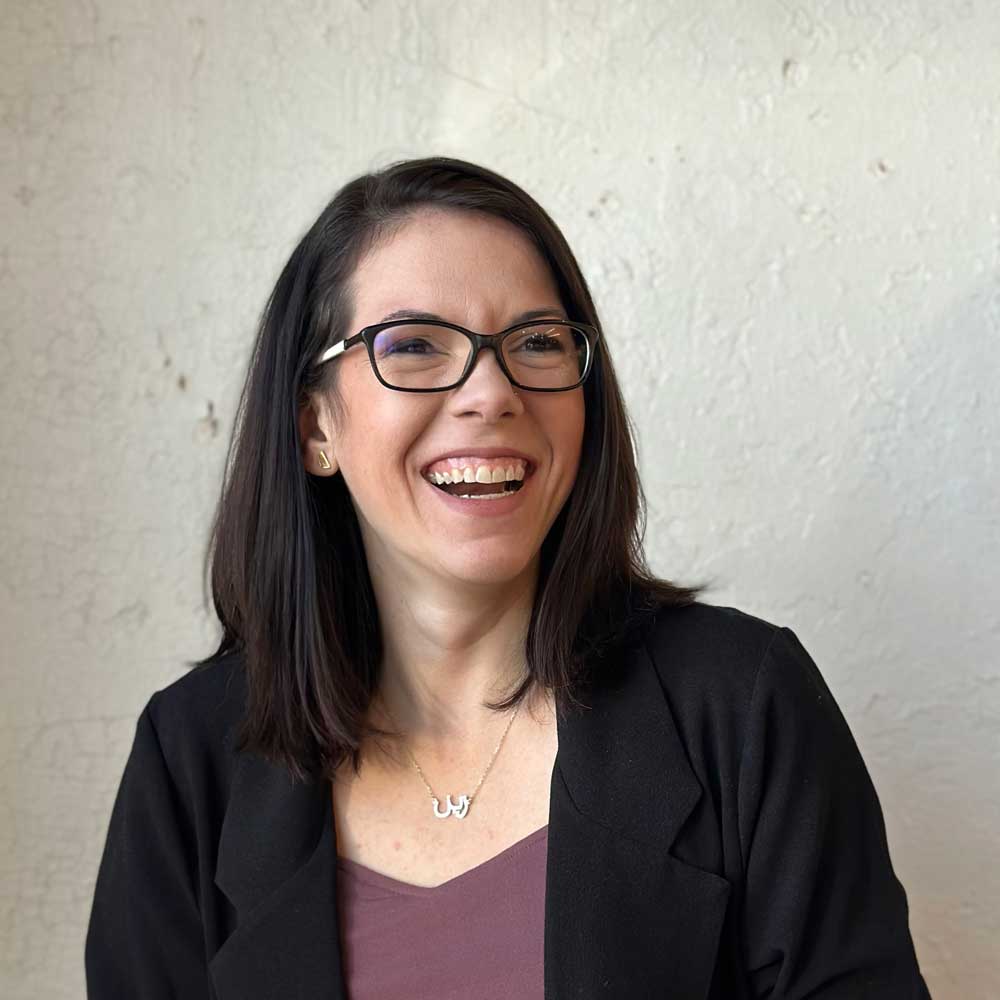 The image shows a smiling person with glasses, wearing a black jacket over a purple top, against a textured beige background. They seem joyful.