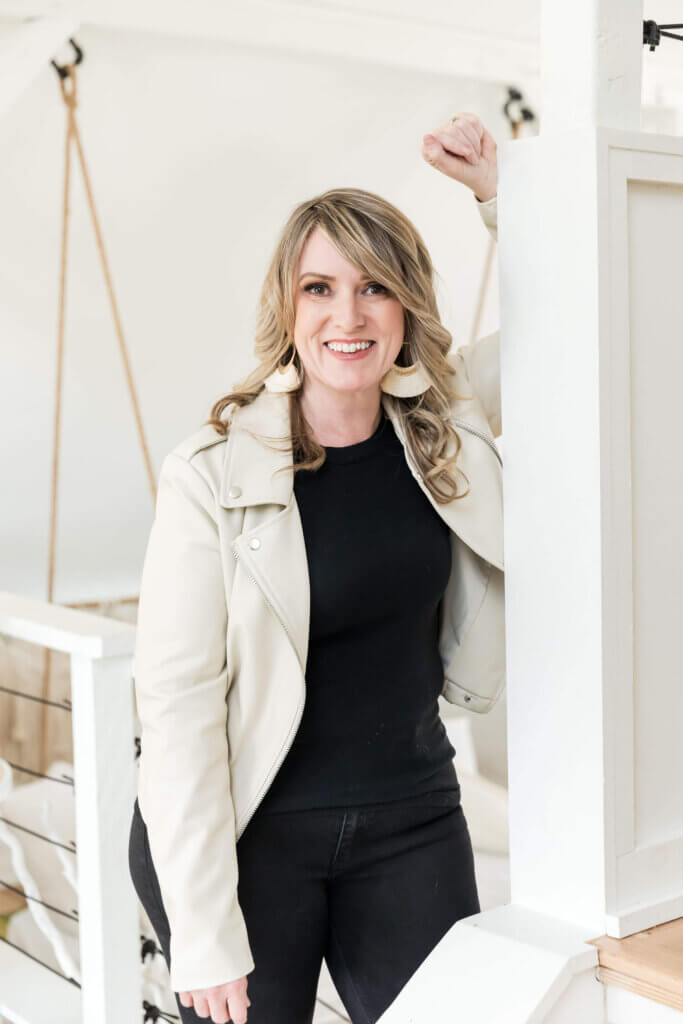 A smiling person leans on a stair railing, wearing a cream jacket, black top, and trousers, with striped earrings in a bright interior.