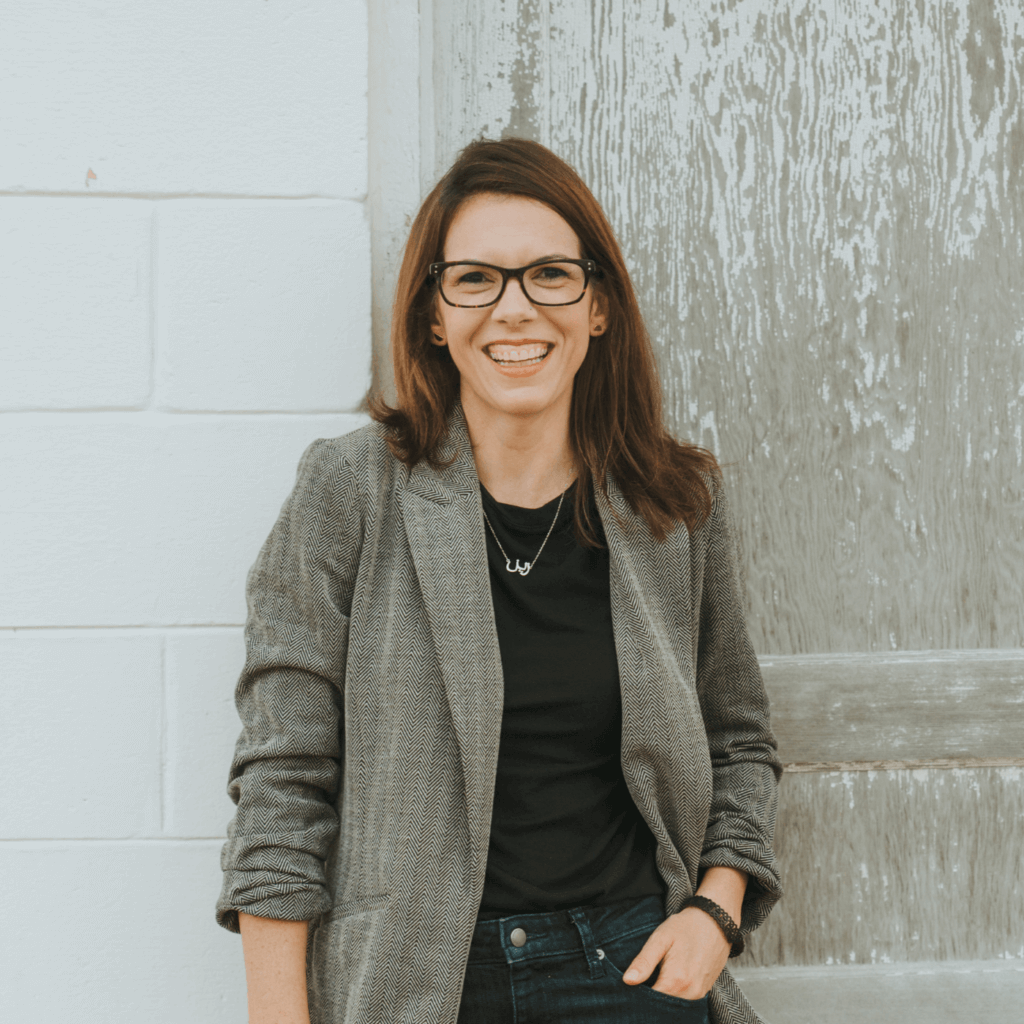 A smiling person with glasses, wearing a blazer, stands against a rustic door and white brick wall background, exuding a cheerful demeanor.