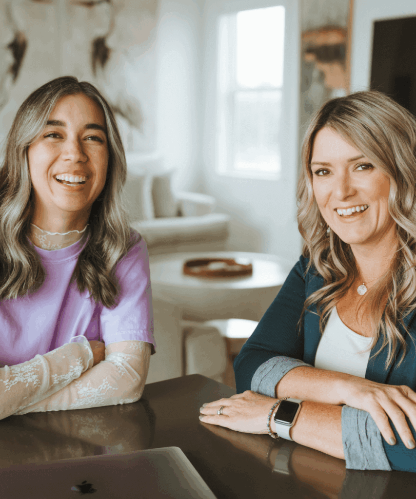 Two smiling people sit at a table in a bright room, with a laptop and art in the background.