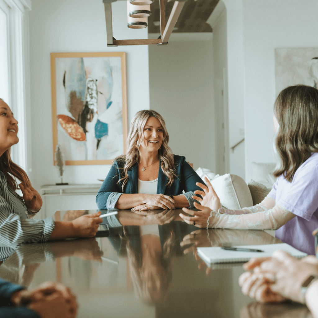 A group of people are having a discussion around a table in a bright room with abstract artwork on the wall.