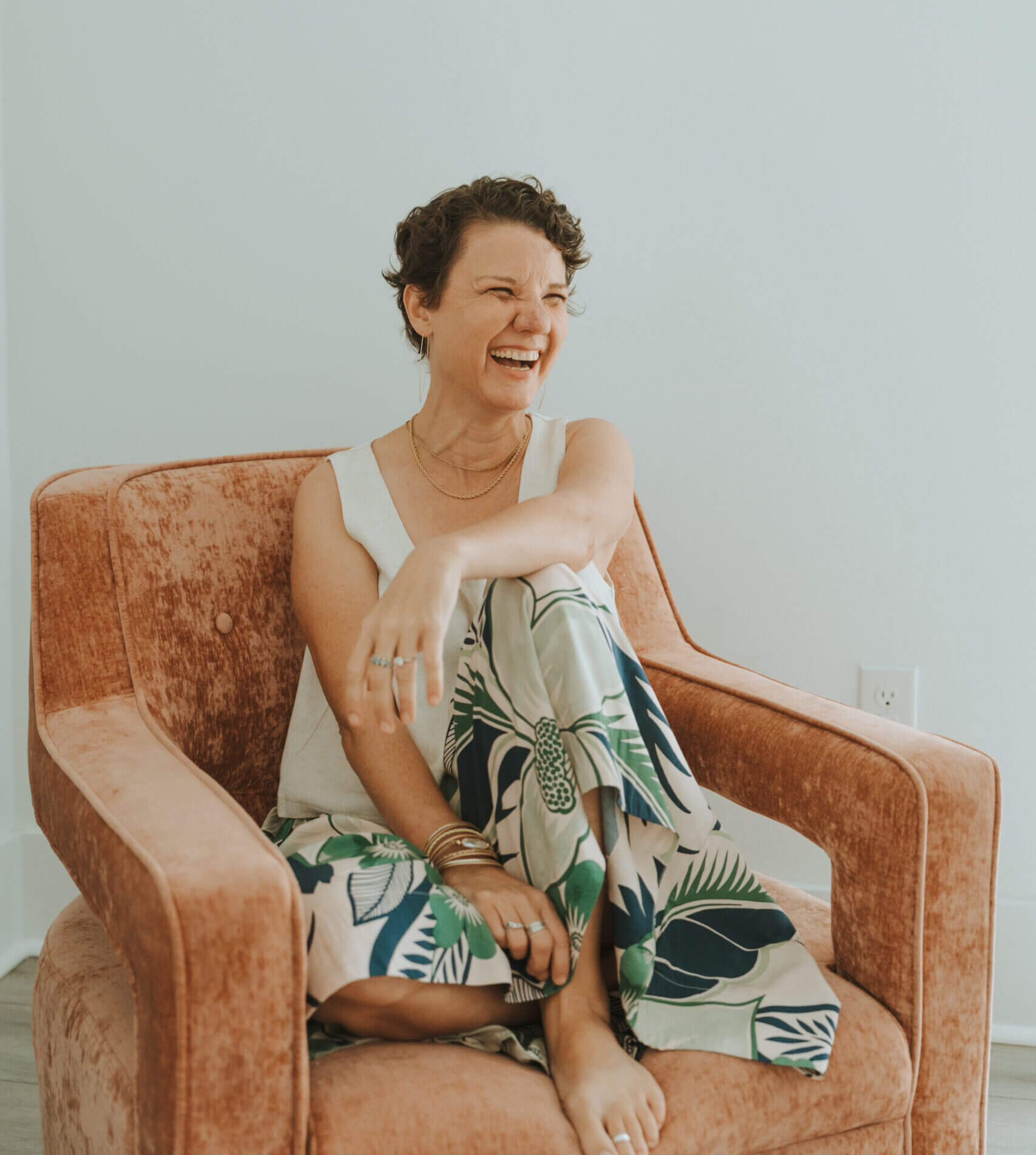 A person sits laughing on a peach-colored chair, wearing patterned pants and a white top. The background is a plain, white wall.