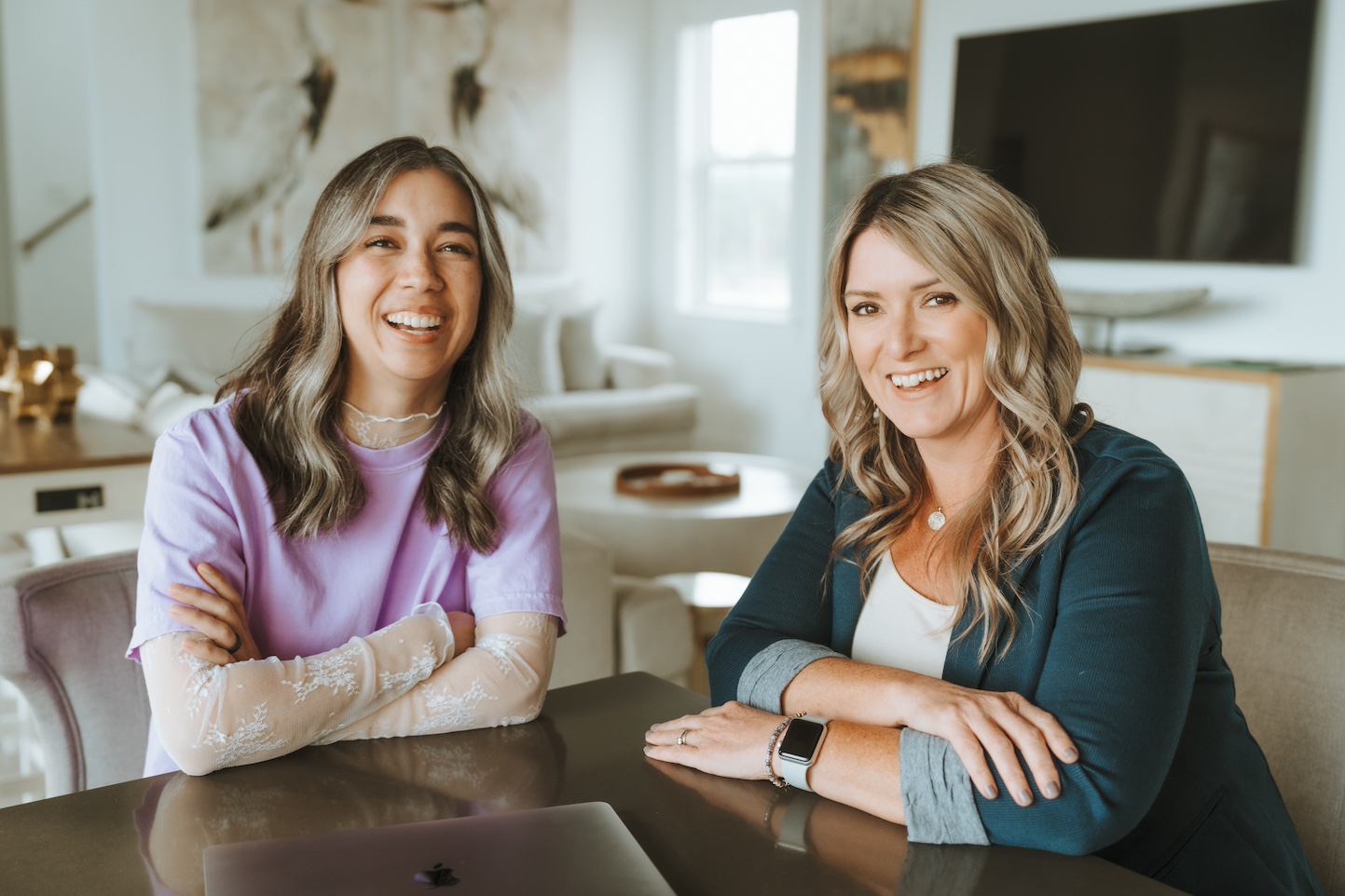 Two smiling people sit at a table in a modern, cozy room with neutral decor and a large TV in the background.