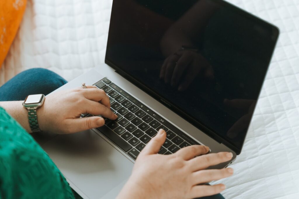 A person in a green top uses a laptop with a dark screen, wearing a green watch. The background is a white quilt.