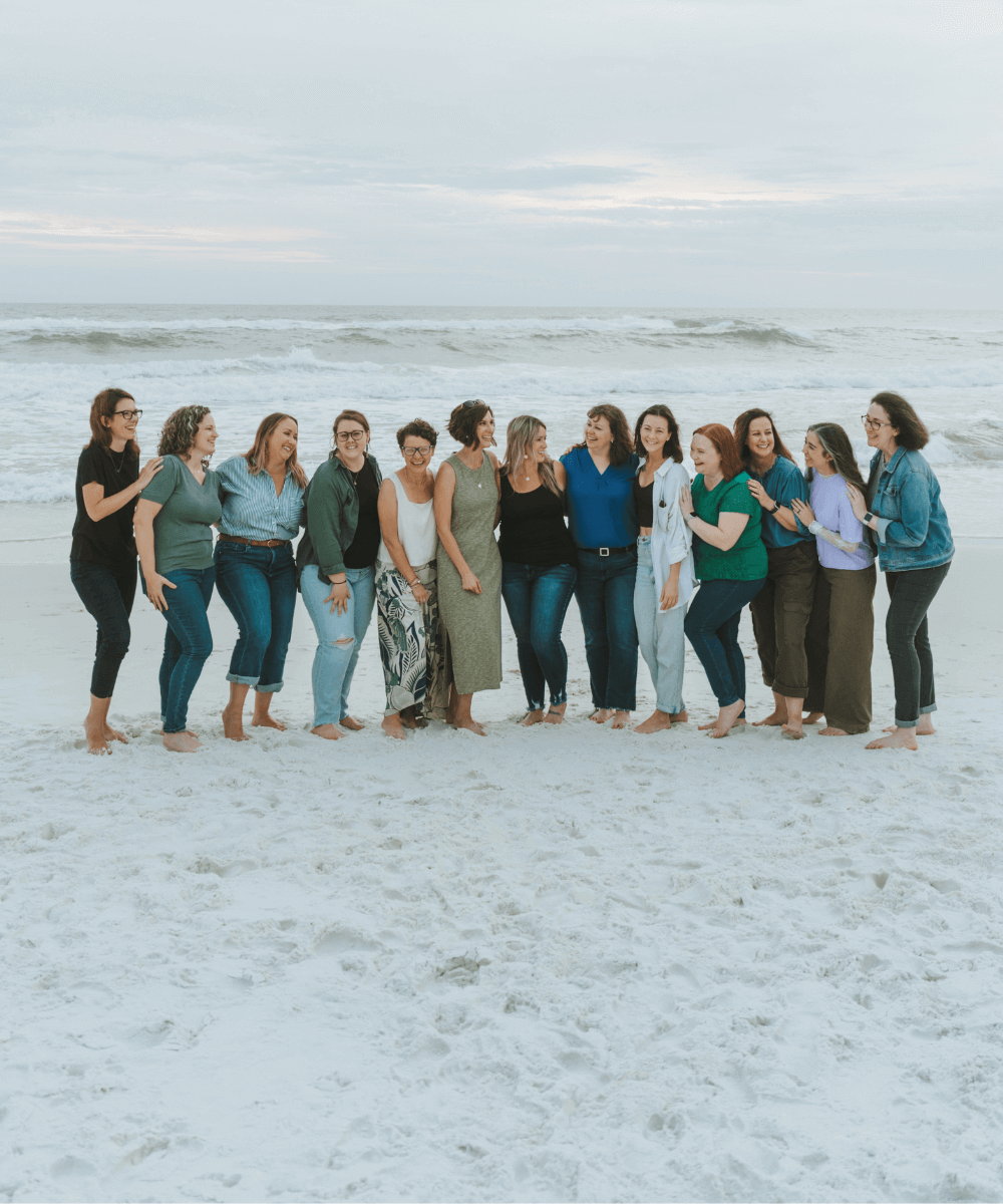 A group of people standing closely together, smiling and laughing on a sandy beach with waves in the background under a cloudy sky.