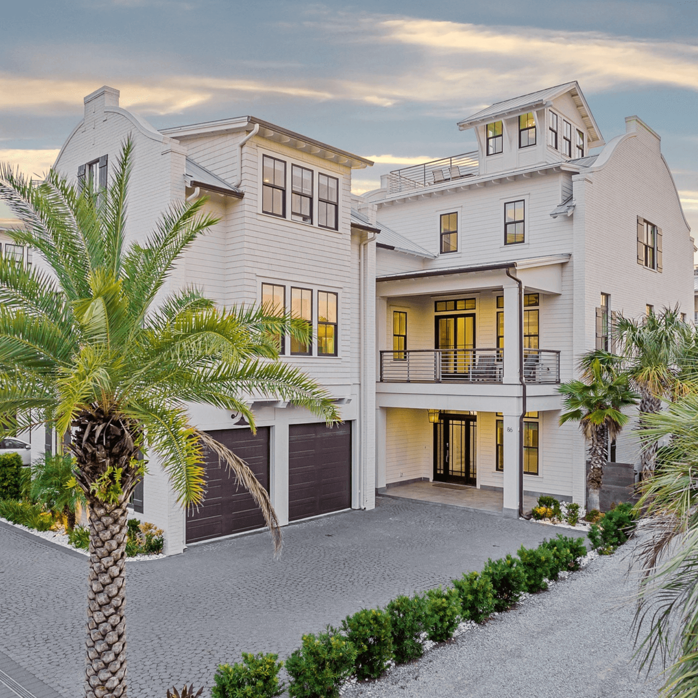 A modern, multi-story white house with palm trees, large windows, and two garage doors under a cloudy sky at sunset.