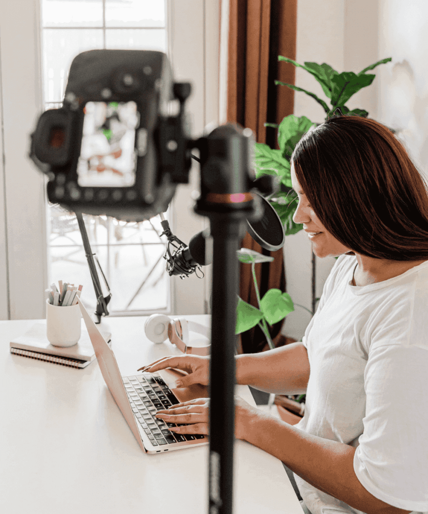 A person types on a laptop in a home office setup. A camera and microphone are positioned nearby, with plants in the background.