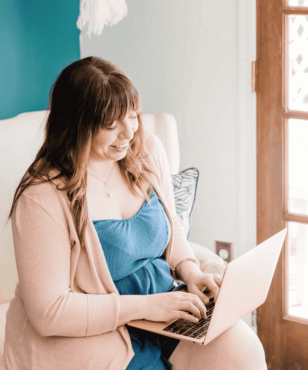 A person using a laptop, sitting in a cozy, well-lit room with blue and beige decor, near a window.
