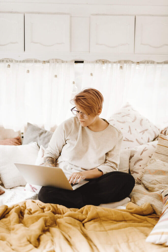 A person with glasses uses a laptop while sitting comfortably on a bed with pillows, in a cozy, bright room.