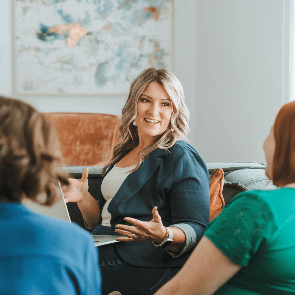 A person with a laptop engages in conversation with two others, sitting in a cozy room with abstract art on the wall.