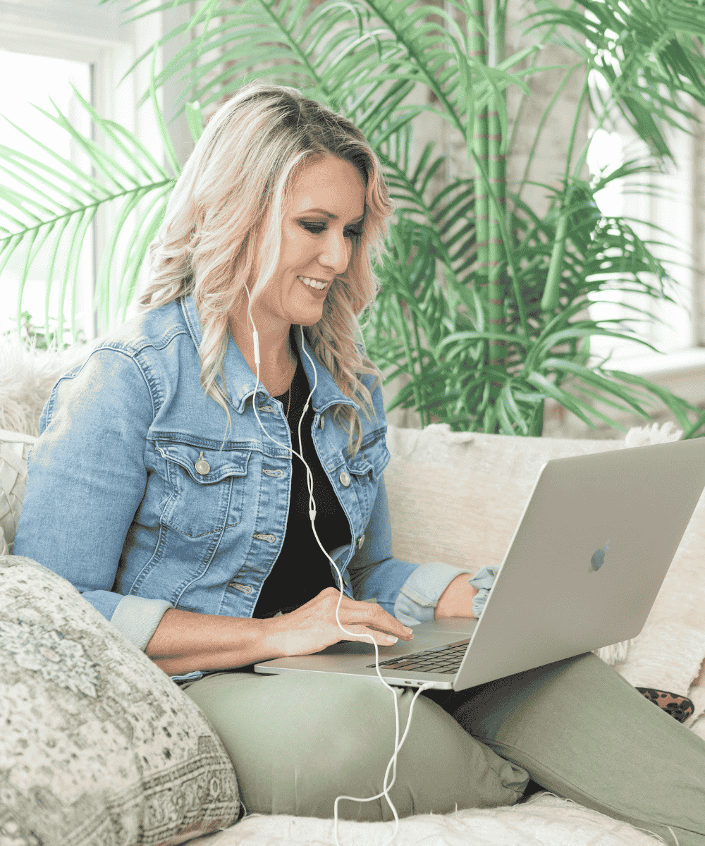 A person with headphones uses a laptop while sitting on a sofa, surrounded by cushions and green plants, in a well-lit room.