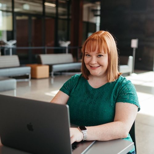 A person in a green shirt smiles, working on a laptop at a modern indoor setting with lounge chairs and reflective glass walls.