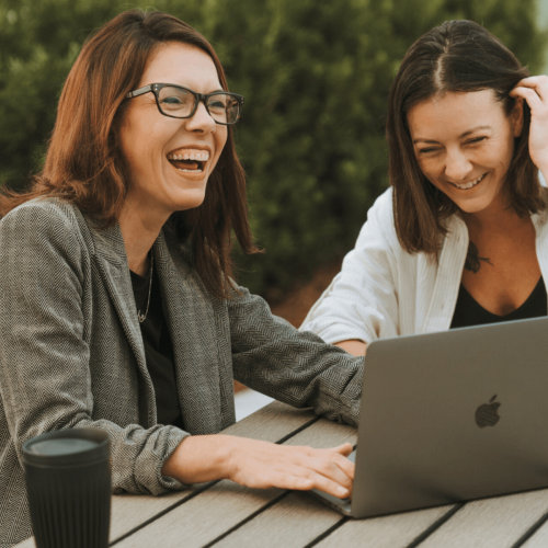 Two people are laughing together while looking at a laptop outside on a wooden table, with greenery in the background.
