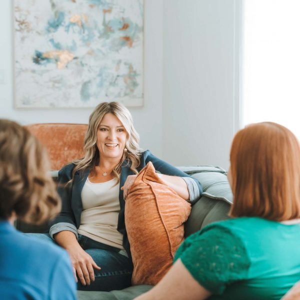 Three people sit on a couch having a conversation. A colorful abstract painting hangs on the wall behind them.