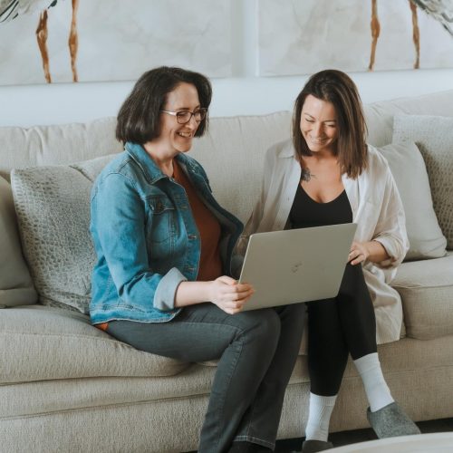 Two people sitting on a sofa, smiling while looking at a laptop. Cozy room with abstract artwork visible in the background.