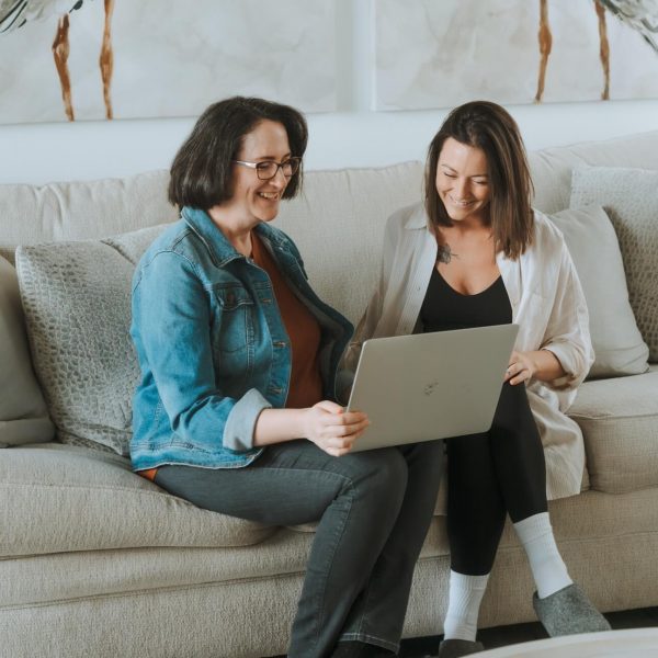 Two people sitting on a sofa, smiling while looking at a laptop. Cozy room with abstract artwork visible in the background.