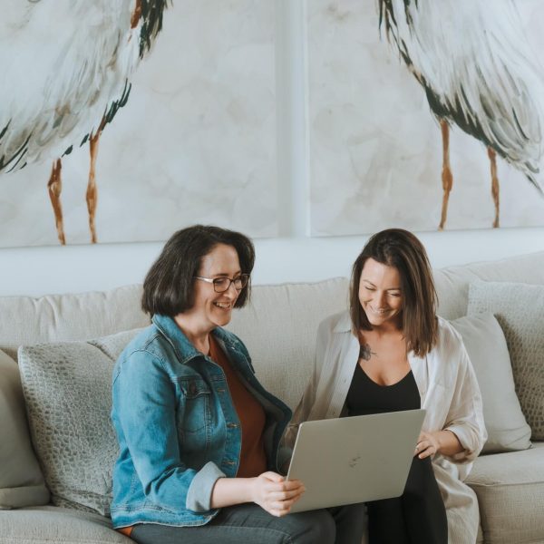 Two people sit on a sofa, smiling while looking at a laptop. Large bird paintings are displayed on the wall behind them.