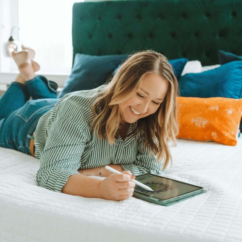A person is lying on a bed, using a tablet with a stylus, surrounded by colorful pillows in a cozy bedroom setting.