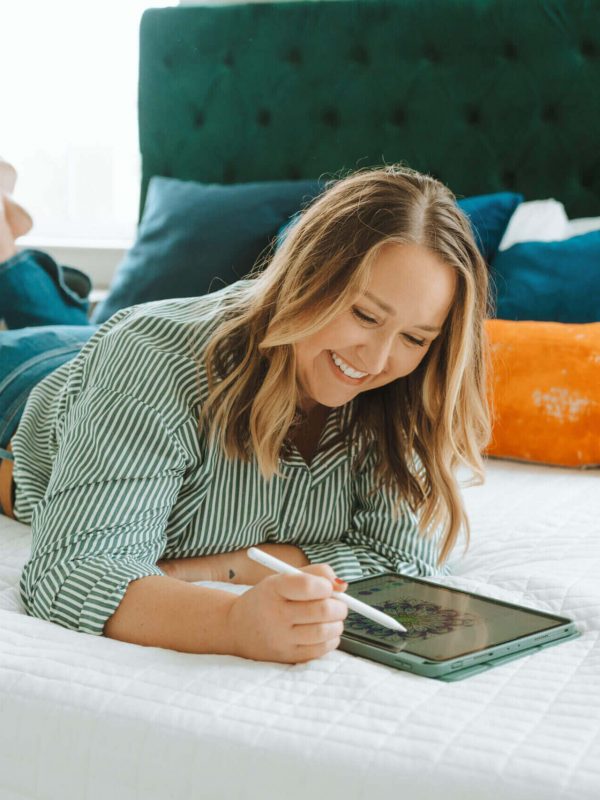 A person is lying on a bed, using a tablet with a stylus, surrounded by colorful pillows in a cozy bedroom setting.