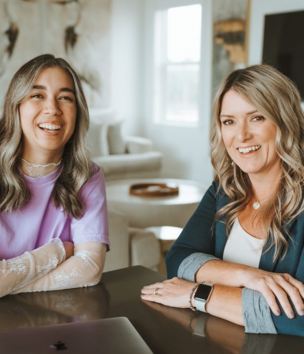 Two smiling people sit at a table in a modern, cozy room with neutral decor and a large TV in the background.