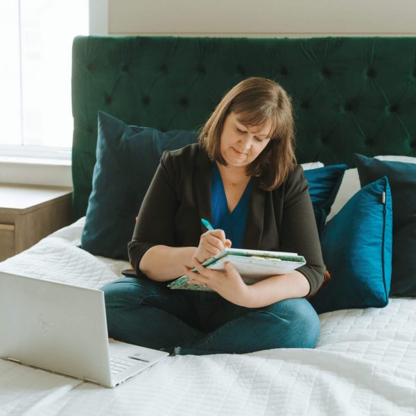 A person sits on a bed, writing in a notebook, with a laptop nearby. The room features a large green headboard.