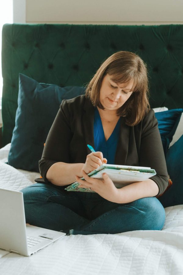 A person sits on a bed, writing in a notebook, with a laptop nearby. The room features a large green headboard.