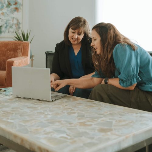 Two people seated at a table, engaging with a laptop. One person is pointing at the screen, in a well-lit room.