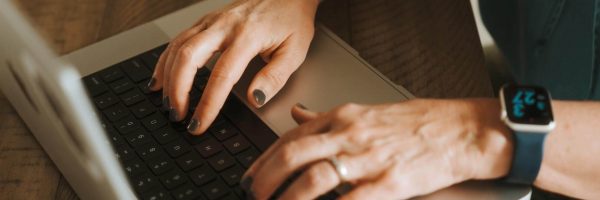 A person typing on a laptop wearing a smartwatch with a blue band, positioned on a wooden surface.
