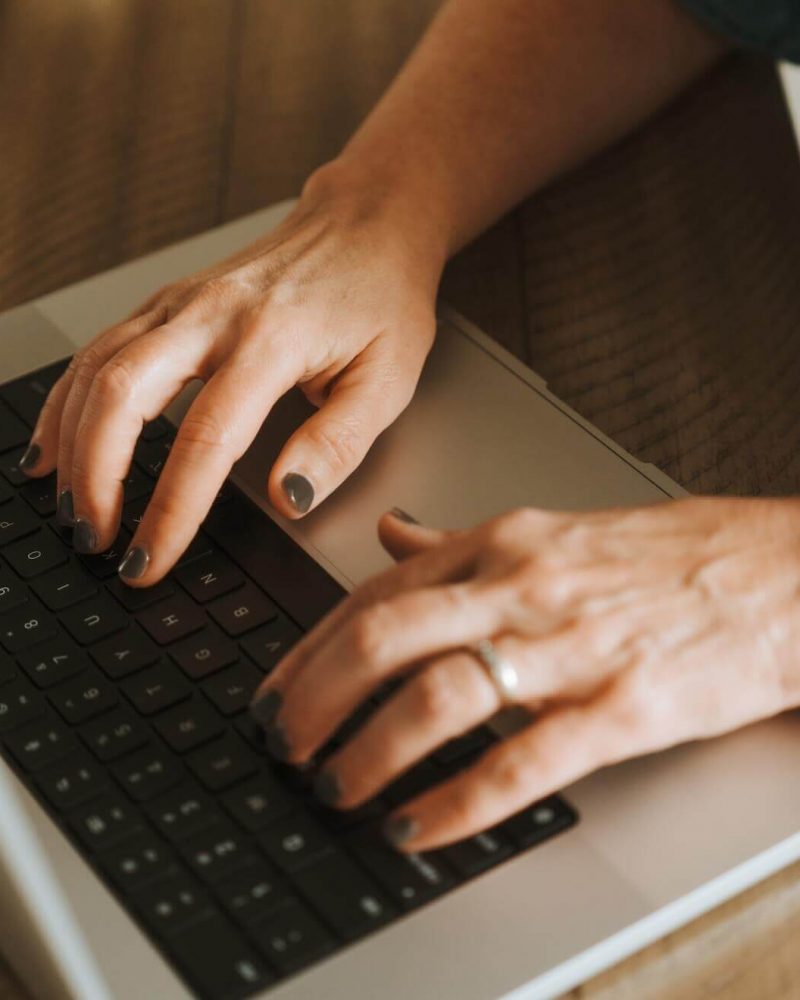 A person typing on a laptop wearing a smartwatch with a blue band, positioned on a wooden surface.