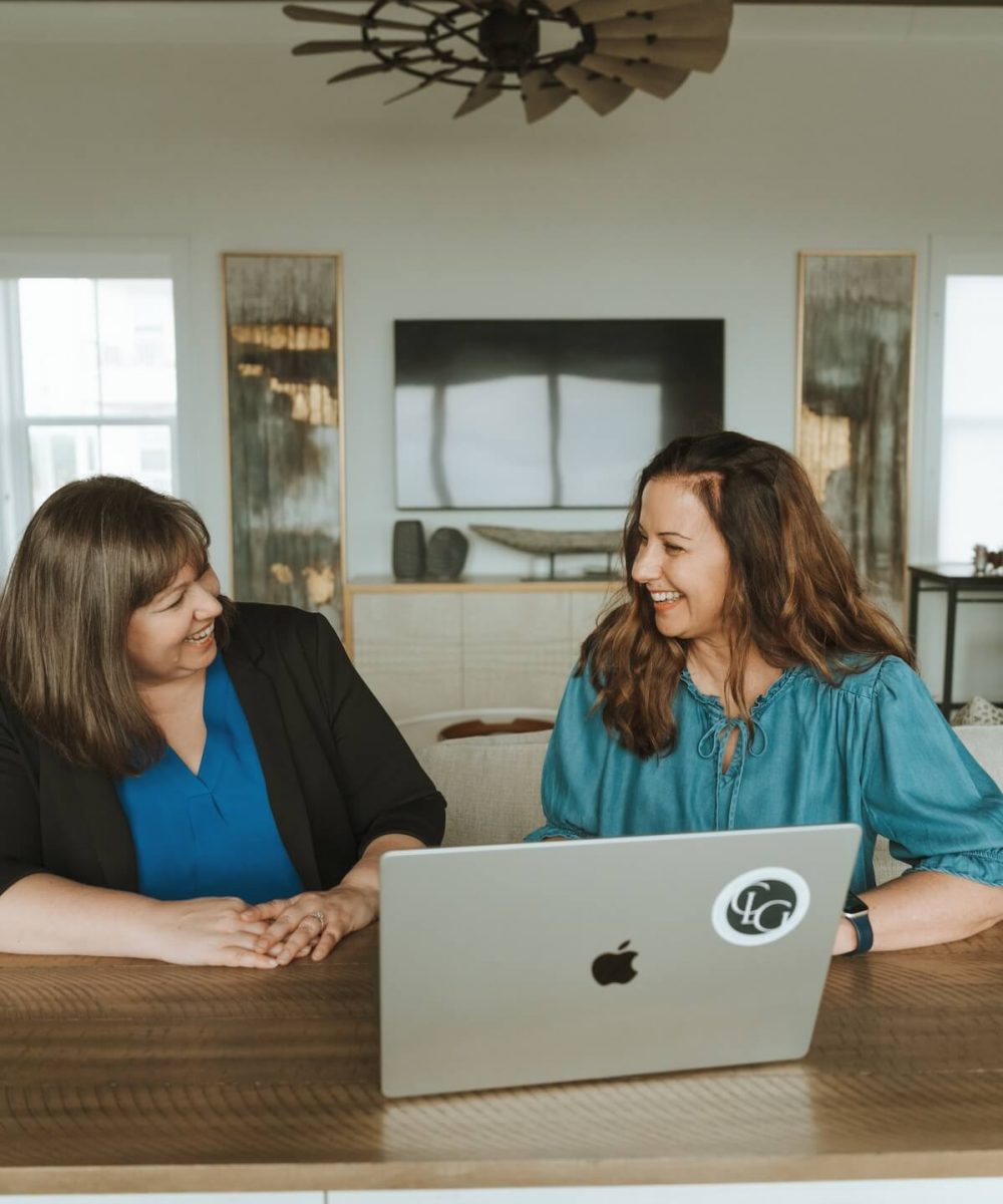 Two people smiling and talking at a wooden table with a laptop. Modern living room decor in the background.
