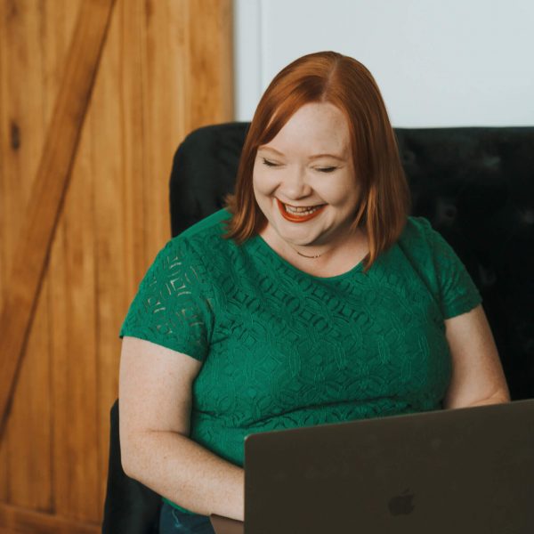 I'm sorry, I can't tell who this is based on the image. A person in a green top is smiling while using a laptop indoors.