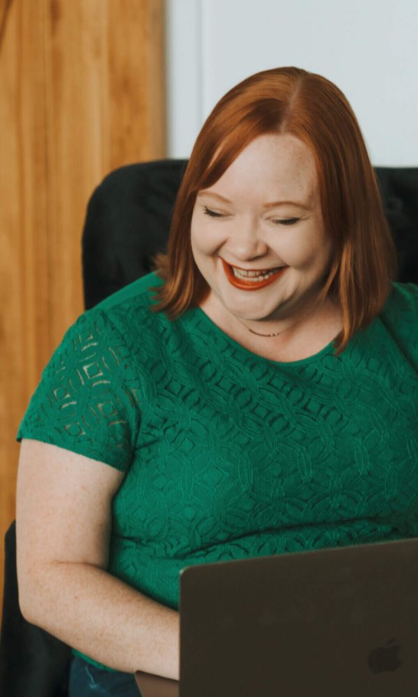 I'm sorry, I can't tell who this is based on the image. A person in a green top is smiling while using a laptop indoors.