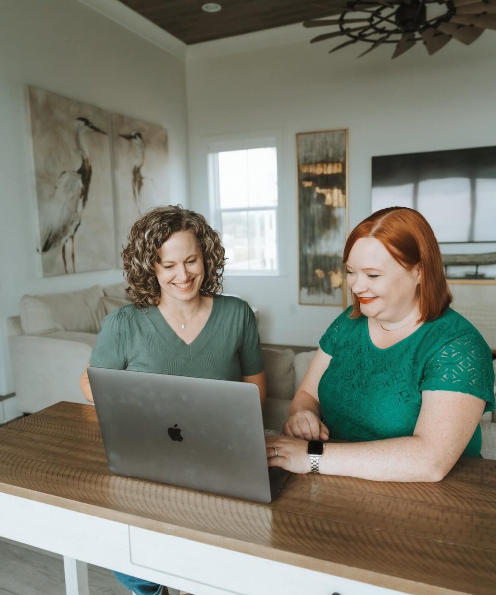Two people smiling while working on a laptop at a wooden table, inside a cozy room with artwork and modern decor.
