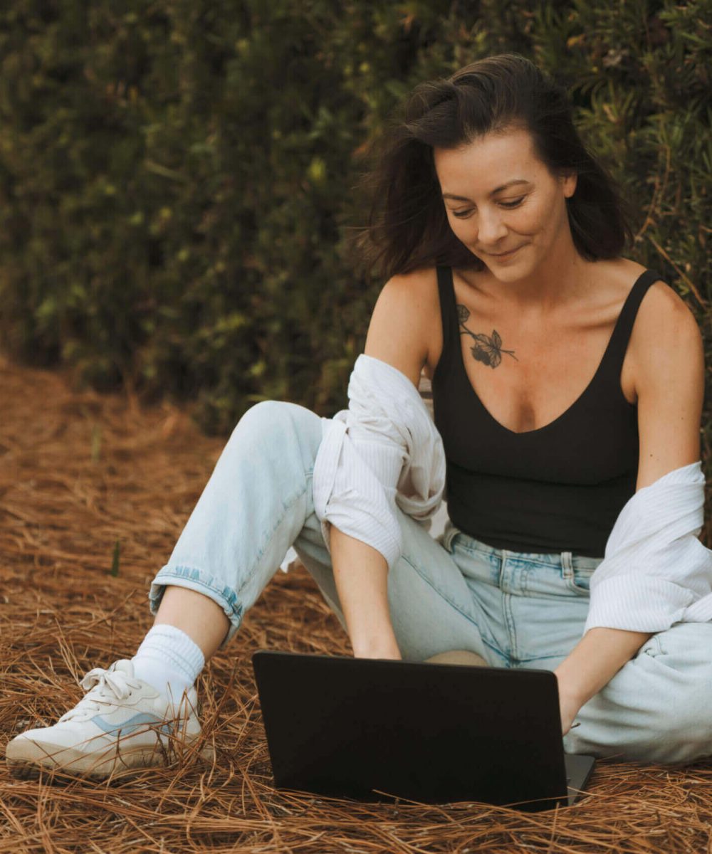 A person sits outdoors on pine needles, using a laptop. They're wearing casual clothing, including a black top and light jeans.