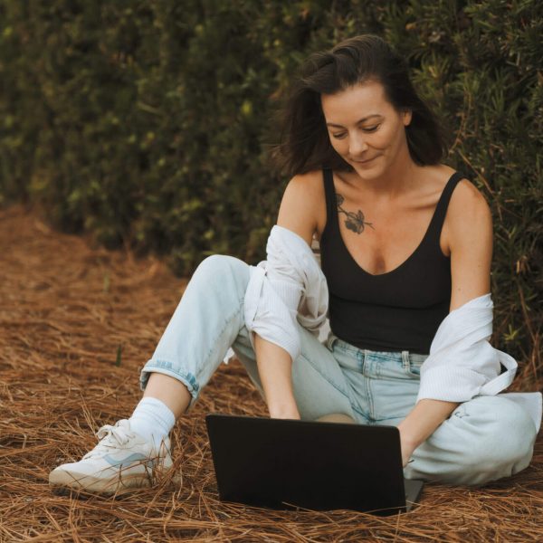 A person sits outdoors on pine needles, using a laptop. They wear casual attire: jeans, a tank top, and a white shirt.