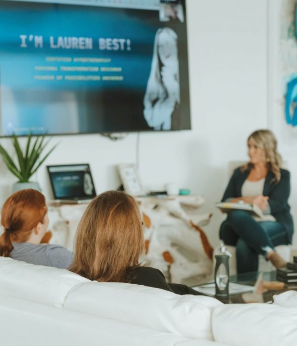 Four people sit on white couches in a modern meeting room, watching a presentation on a wall-mounted screen.