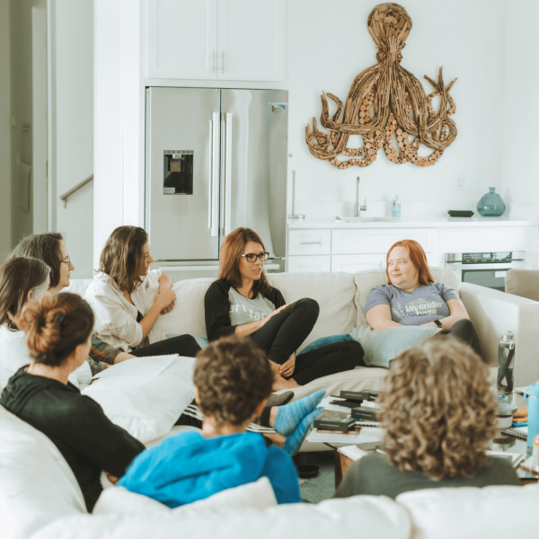 A group of seven people sits in a cozy living room, engaged in conversation, with wooden octopus wall art and modern decor.