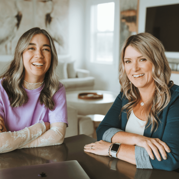 Two people smiling at a table in a bright room with art, a couch, and a window in the background.