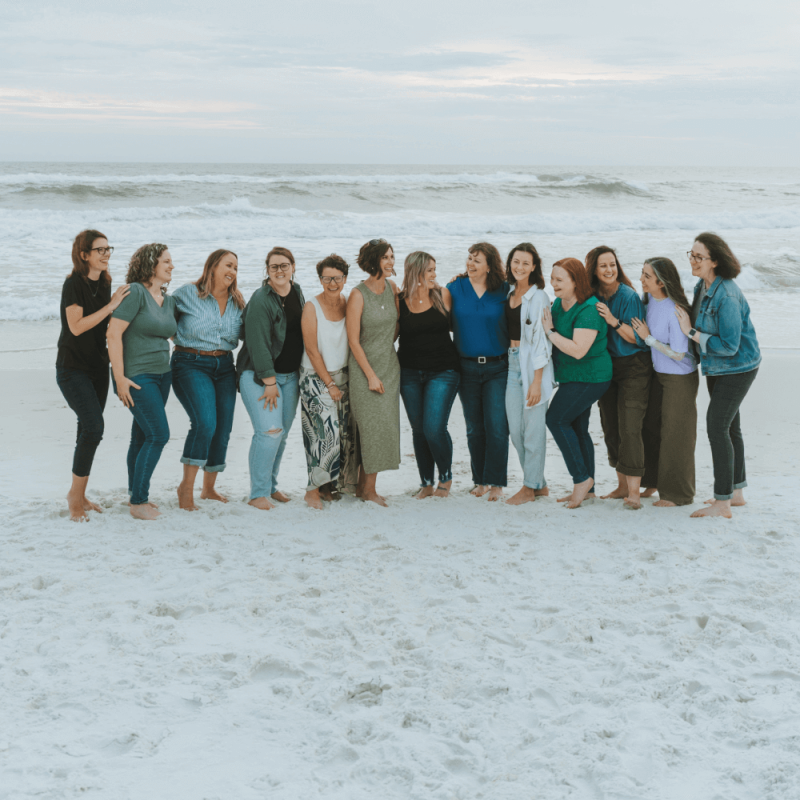 A group of people standing closely together, smiling and laughing on a sandy beach with waves in the background under a cloudy sky.