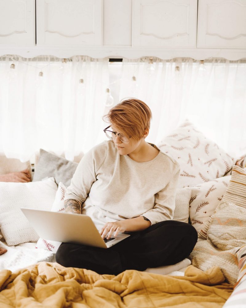 A person with short hair and glasses sits comfortably on a bed, typing on a laptop in a cozy, well-lit room.